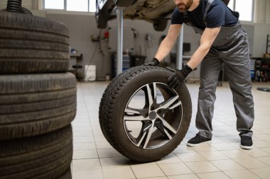 Profesyonel oto tamirhanesinde tamirci araba lastiği değiştiriyor. Tamir ve onarım için lastik taşıyan üniformalı bir işçi. Otomotiv hizmeti kavramı garaj ortamında kaydedildi.