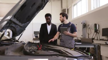 İş yerindeki tamirci, modern oto tamirhanesinde takım elbiseli müşterilere araba sorunlarını anlatıyor. Araç bakım ve onarım süreciyle ilgili profesyonel danışma.