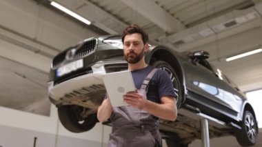 Tamirci, kaldırılmış aracın altındaki araba tamirhanesinde tablet kullanıyor. Odaklanmış otomotiv teknisyeni teşhisi inceliyor, modern otomobil bakımı teknolojisi, profesyonel hizmet ortamı.