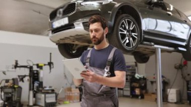 Dost canlısı tamirci, modern araba tamirhanesinde tablet kullanıyor ve asansörde araba var. Profesyonel teknisyen araç kontrolü ve bakımını kontrol ediyor. Otomotiv endüstrisi ve müşteri hizmetleri kavramı.