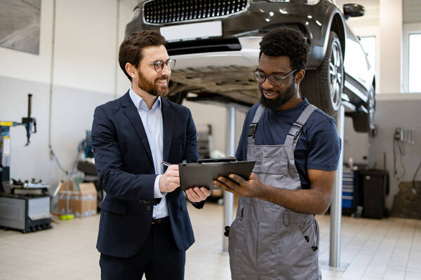 Механик и бизнесмен в костюме обсуждают обслуживание автомобилей в гараже. Человек в комбинезоне держит планшет, улыбается. Профессиональное взаимодействие, автомобильный сервис.