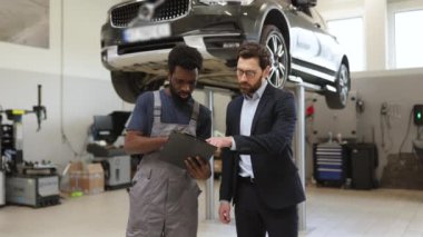 Tamirci modern tamirhanedeki müşteriye araba tamir detaylarını anlatıyor. Araç hizmet merkezinde tamirci ve istemci arasında asansörde araba ile profesyonel etkileşim.