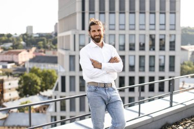 Beyaz gömlekli ve kot pantolonlu kendine güvenen işadamı çatıda kolları bağlı ve arka planda şehir manzarası var. Başarı kavramı, kentsel yaşam tarzı ve profesyonel güven.