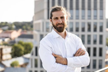 Sakallı, uzun saçlı, beyaz gömlekli, kollarını kavuşturmuş kendine güvenen bir iş adamı. Arka planında modern yüksek binalar olan şehir manzarası.