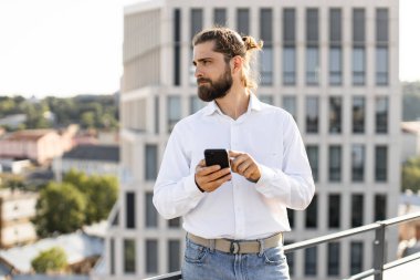 Beyaz gömlekli bir adam arka planda şehir manzarası olan akıllı telefon kullanıyor. Profesyonel iş adamı dışarıdaki telefonları kontrol ediyor. Gökyüzü açık modern ofis binası.