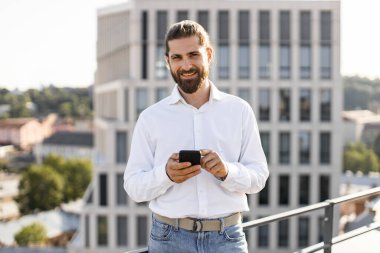 Modern ofis binasının çatısında akıllı telefon kullanarak sakallı gülümseyen adam. Beyaz gömlek ve kot pantolon giyen bir işadamı kentin arka planına karşı duruyor. Kendinden emin ve nişanlı görünüyor..