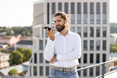 Beyaz tişörtlü sakallı adam şehir merkezindeki çatıda dururken akıllı telefondan konuşuyor. Arka planda ofis binaları olan şehir merkezi. Güneşli bir günde açık havada sıradan iş konuşmaları..