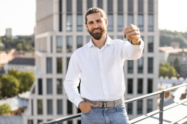 Modern binanın çatısında anahtarlarını tutan neşeli genç adam yeni daireye taşınmamızı kutluyor. Başarı kavramı ve yeni başlangıçlar.