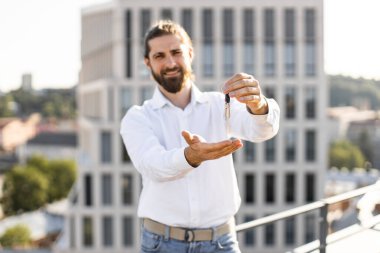Beyaz tişörtlü sakallı adam elinde arka planda binalar olan anahtarlar tutuyor. Gayrimenkul ve ev sahipliği kavramı