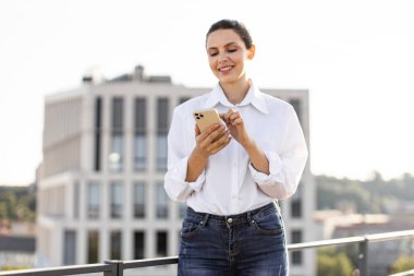 Sıradan kıyafetlerle gülümseyen kadın çatıda akıllı telefon kullanıyor ve şehir geçmişi bulanık. Modern iletişim, teknoloji ve sosyal medya kavramı.