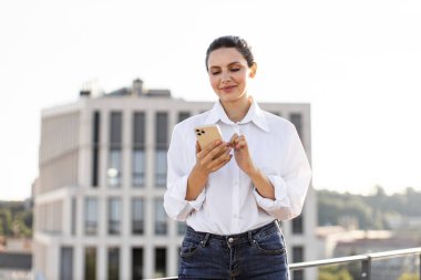 Çatıda duran kadın akıllı telefon kullanıyor, beyaz gömlek ve kot pantolon giyiyor, arka planda modern şehir manzarası var. Cep telefonunu kontrol ederken odaklanmış ve mutlu görünüyor..