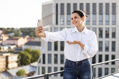 Gülümseyen kadın arka planda manzarası olan balkonda selfie çekiyor. Günlük kıyafet, rahat atmosfer ve güzel bir gün..