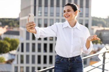 Arka planda şehir binalarıyla çatıda selfie çeken mutlu kadın. Güneşli açık hava ortamının keyfini çıkarırken, gülümseyen ve telefonla meşgul olan biri..