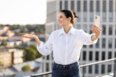 Çatıda duran bir kadın, arka planda şehir manzarasını yakalayan akıllı telefonuyla selfie çekiyor. Beyaz gömlek ve kot pantolon giyip güneşli bir günde dışarıda eğlenmek..