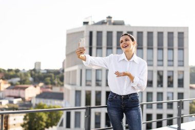 Sıradan kıyafetlerle gülümseyen kadın çatıdaki terasta selfie çekiyor. Açık havada, şehir binaları ve arka planda açık gökyüzü ile neşeli bir anı yakalıyor..