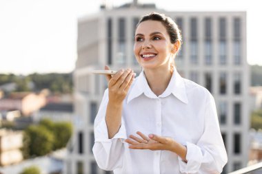 Şehir ortamında akıllı telefon ses asistanı kullanarak gülümseyen bir iş kadını. Arka planda modern bina olan açık havada duran beyaz gömlekli bir kadın. Teknoloji iletişim işi kavramı.