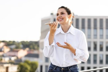 Çatının terasında arka planda akıllı telefon asistanı kullanan gülümseyen bir kadın. Günlük beyaz gömlek ve kot giyen kadın gündüz iletişiminin keyfini çıkarıyor..