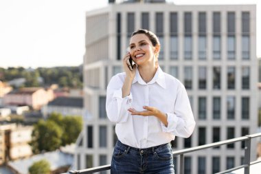Çatıda durmuş gülümseyen bir kadın cep telefonuyla konuşuyor ve şehir manzarasının tadını çıkarıyor. Beyaz gömlekli, kot pantolonlu, rahat ve profesyonel tarzlı..