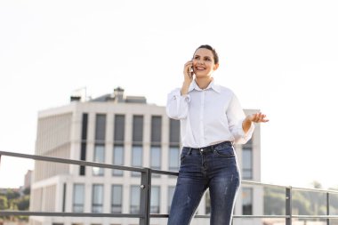 Modern ofis binasının dışında cep telefonuyla konuşan kendinden emin bir kadın. Mutlu görünüyor, rahat, günlük kıyafetler giyiyor, parlak gün ışığı, başarılı bir mesleki hayatı olduğunu gösteriyor..