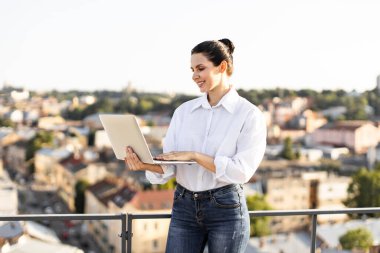 Çatıda dizüstü bilgisayar kullanan gülümseyen bir kadın. Uzaktan çalışan profesyonel, şehir ortamının tadını çıkaran. Günlük giysiler içinde beyaz kadın, ekrana odaklanmış..
