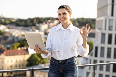 Çatıda dizüstü bilgisayarı tutan kendine güvenen bir kadın tamam işareti veriyor. Profesyonel kıyafet, olumlu ifade, arka planda şehir manzarası. Başarılı iş konsepti.
