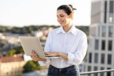 Şehir ortamında dizüstü bilgisayar kullanan genç bir kadın gülümsüyor ve mutlu görünüyor. Günlük kıyafetler rahat ama üretken bir ortam olduğunu gösteriyor. Arkaplanda modern binalar ve şehir manzarası yer alıyor.