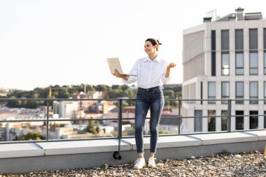 Gülümseyen kadın elinde dizüstü bilgisayarla çatıda duruyor, şehir manzarasının tadını çıkarıyor ve uzaktan çalışıyor. Profesyonel ve sıradan, modern çalışma esnekliği ve yaşam tarzını somutlaştırıyor..