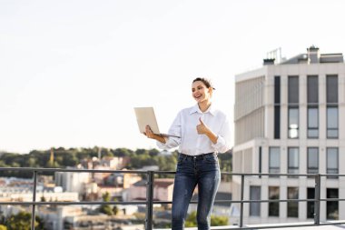 Laptoplu kendine güvenen genç bir kadın şehir binalarının arka planında, açık terasta başparmak kaldırıyor. Güneşli havada günlük kıyafetler, başarıyı ifade eder, başarıyı..