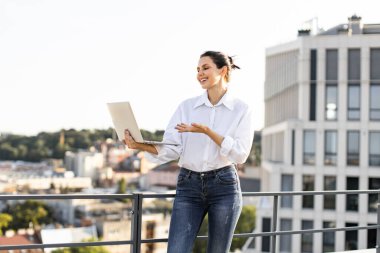 Çatıdaki terasta dizüstü bilgisayar kullanan gülümseyen kadın. Uzaktan çalışma, teknoloji ve şehir yaşam tarzı kavramı. Kadın çalışırken temiz havanın tadını çıkarıyor. İş kadını çevrimiçi toplantı düzenliyor.