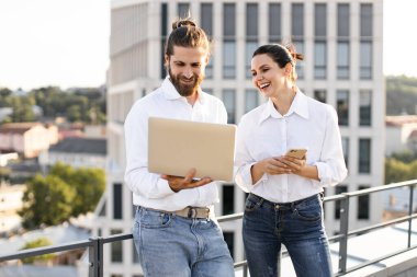 Çatıda dizüstü bilgisayar ve telefon kullanarak proje tartışırken gülümseyen iki iş arkadaşı, günlük beyaz gömlekler ve kot pantolon giyiyor, kentsel arka plan.