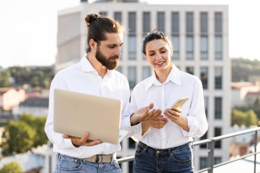 Açık şehir ortamında dizüstü bilgisayar ve akıllı telefon kullanırken iş tartışan genç profesyoneller. Takım çalışması, işbirliği ve teknolojiden anlayan bir yaklaşım. Arka planda şehir manzarası.