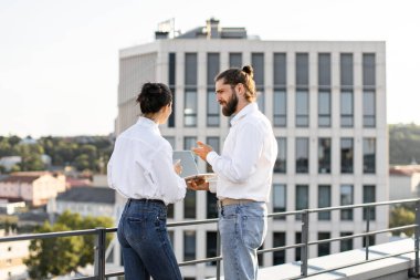 İki meslektaş, arka planda şehir silueti ve dizüstü bilgisayarla çatıda çalışmayı tartışıyor. Günlük iş ortamı, iletişim ve işbirliğine odaklan.