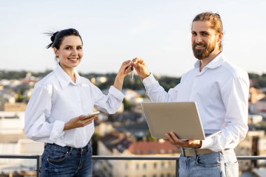 Şehrin manzaralı çatıda yeni ev alımını kutlayan gülümseyen bir çift. Kadın anahtarları tutuyor, adam dizüstü bilgisayar tutuyor. Neşeli ve heyecanlı an yeni başlangıçları ve gelecekteki hedefleri simgeliyor..