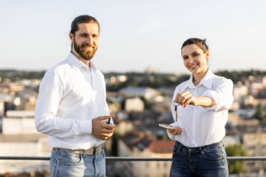 Çatıda duran gülümseyen çift şehrin arka planında anahtarlarını tutuyor. Gayrimenkul, yeni ev sahipliği, başarı ve mutluluk kavramı. Parlak güneşli bir gün Neşeli bir ruh hali yaratıyor.
