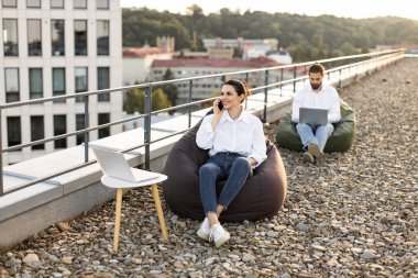 Çatıda dizüstü bilgisayar kullanarak çalışan iş adamları, temiz havanın ve alternatif çalışma alanının tadını çıkarıyorlar. Günlük kıyafetler, rahat ortam, modern çalışma alanı kavramları.