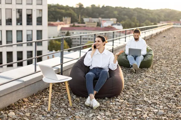 İş arkadaşları çatıda dizüstü bilgisayarlarla çalışıyor, açık havada eğleniyorlar. Telefondaki kadın, dizüstü bilgisayara odaklanmış bir adam, ikisi de sıradan giyinmiş. Uzak çalışma ve açık hava çalışma alanı kavramı.