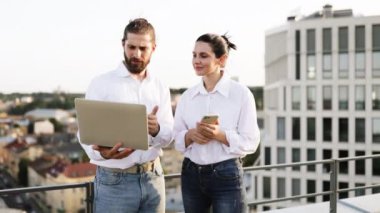 Çatıda dizüstü bilgisayar ve telefonla iş tartışan genç profesyoneller. Şehir ortamında rahat bir iş ortamı. Takım çalışması için mükemmel, modern çalışma stilleri.