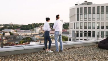 İki meslektaş, arka planda şehir silueti ve dizüstü bilgisayarla çatıda çalışmayı tartışıyor. Günlük iş ortamı, iletişim ve işbirliğine odaklan.