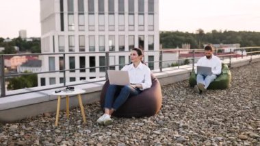 Profesyoneller, laptoplarda, açık hava ortamının tadını çıkararak çatıdaki günlük iş yerlerinde, fasulye torbalarıyla çalışıyorlar. Ortak çalışma ayarları modern iş ve uzak çalışma eğilimlerini yansıtır.