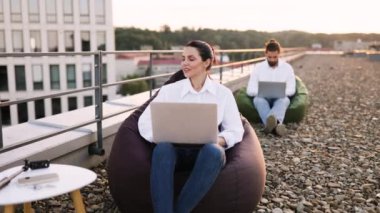 Ofisin çatısındaki fasulye çuvallarında otururken dizüstü bilgisayarlarda çalışan iş adamları. Açık havada rahat çalışma ortamı, esnek çalışma alanı kavramı.