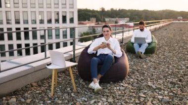 Çatıda uzaktan çalışmaktan zevk alan iki kişi. Adam dizüstü bilgisayarda çalışıyor, kadın fasulye torbalarının üzerinde otururken akıllı telefon kullanıyor. Manzaralı bir çalışma alanı düzeneği. Rastgele çalışma ortamı.