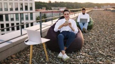 Laptop ve akıllı telefon kullanan iki kişinin oturduğu sıradan bir çatı çalışma ortamı. Rahat ve verimli çalışma alanı sahnesi. Modern ofis binası şehir ortamını geliştirdi