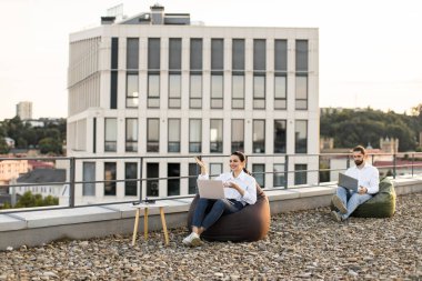Modern bir ofis binasının önünde dizüstü bilgisayar kullanarak çatıda uzaktan çalışan iki profesyonel. Açık havada oturma, esnek ve yenilikçi çalışma ortamları ile rahat çalışma ortamı.