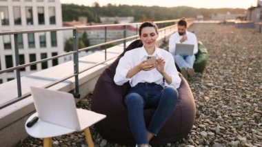 Çatıdaki terasta oturan genç bir kadın akıllı telefon kullanıyor ve başparmağını kaldırıyor. Rahat ve rahat bir atmosfer, rahat fasulye şezlongunda dışarıda oturmanın tadını çıkarıyor. Mutlu ve içerik ifadesi.