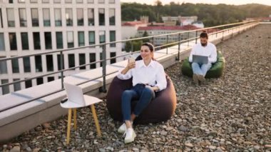 Çatıda uzaktan çalışan iş adamları, dizüstü bilgisayar ve akıllı telefon kullanıyorlar, açık ofis düzenlemelerinin tadını çıkarıyorlar. Fasulye torbaları, iletişim ve teknoloji ile şehir ortamında rahat çalışma ortamı