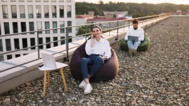 İnsanlar çatıda dizüstü bilgisayar ve fasulye torbaları kullanarak uzaktan çalışıyor. Adam dizüstü bilgisayara odaklanmış, kadın telefonla konuşuyor. Şehir planına sahip rahat bir açık hava çalışma ortamı