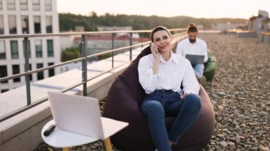 Çatıda uzaktan çalışan iki meslektaş dizüstü bilgisayar ve telefon kullanıyor. Günlük çalışma ortamı gevşemeyi ve üretkenliği teşvik eder. Açık hava düzeni esnekliği ve modern şirket kültürünü ön plana çıkarır