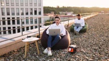 İş kadını koltukta oturuyor ve kabloyu evin çatısındaki taşınabilir elektrik santraline bağlıyor. Arka plandaki meslektaşlar da dizüstü bilgisayarda çalışıyor, verimli bir açık hava çalışma alanı yaratıyor.