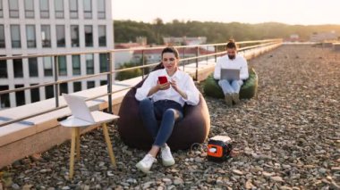 İş adamları çatıdaki puf minderlerde oturup dizüstü bilgisayarlar ve telefonlarla çalışıyorlar. Şarj cihazları için kullanılan taşınabilir güç istasyonu. Kentsel arkaplanı olan rahat bir açık hava çalışma ortamı.