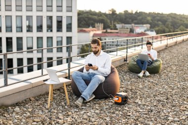 İş adamı puf koltukta oturuyor ve telefon kablolarını evin çatısındaki taşınabilir elektrik santraline bağlıyor. Arka planda dizüstü bilgisayarda çalışan başka biri.